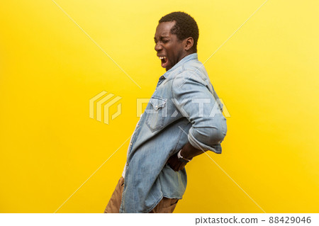 Portrait of unhealthy young man in denim casual shirt holding hands on back with painful grimace, suffering from backache or kidney inflammation. indoor studio shot isolated on yellow background Portrait of unhealthy young man in denim casual shirt holding hands on back with painful grimace, suffering from backache or kidney inflammation. indoor studio shot isolated on yellow background 88429046
