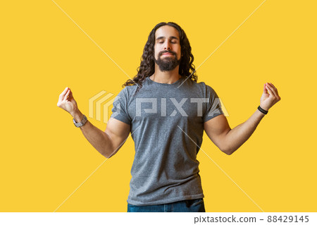 Yoga time. Portrait of calm serious bearded young man with long curly hair in grey tshirt standing in yoga pose and meditating with closed eyes and relaxed. studio shot isolated on yellow background. 88429145