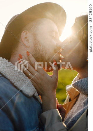 Man and woman in fashion clothes resting on a nature together in sunset. Man wearing jacket and a black hat and woman coat. Photo of romantic couple kissing. Man and woman in fashion clothes resting on a nature together in sunset. Man wearing jacket and a black hat and woman coat. Photo of romantic couple kissing. 88429160