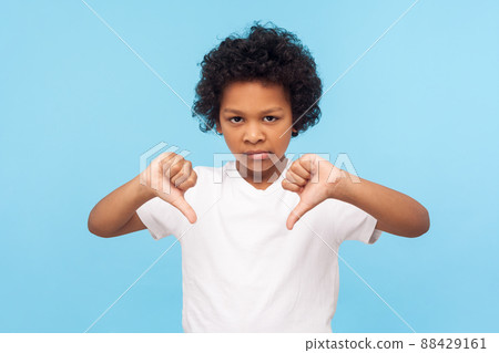 Dislike. Portrait of dissatisfied upset little boy with curly hair showing thumbs down gesture and looking displeased, naughty child making disagree disapprove finger sign. studio shot blue background Dislike. Portrait of dissatisfied upset little boy with curly hair showing thumbs down gesture and looking displeased, naughty child making disagree disapprove finger sign. studio shot blue background 88429161