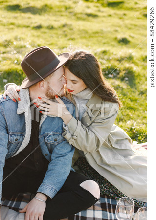 Man and woman in fashion clothes sitting on a nature on a picnic rug. Man wearing jacket and a black hat and woman coat. Woman hugging a man. Man and woman in fashion clothes sitting on a nature on a picnic rug. Man wearing jacket and a black hat and woman coat. Woman hugging a man. 88429266