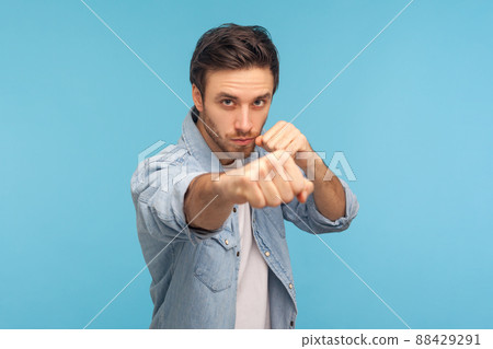 Portrait of man in worker denim shirt standing with boxing gesture, punching to camera, provoking fight or ready to self-defense, struggle concept. indoor studio shot isolated on blue background 88429291