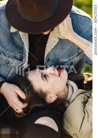 Man and woman in fashion clothes sitting on a nature on a picnic rug. Man wearing jacket and a black hat and woman coat. Woman lying on a man's lap. Man and woman in fashion clothes sitting on a nature on a picnic rug. Man wearing jacket and a black hat and woman coat. Woman lying on a man's lap. 88429303