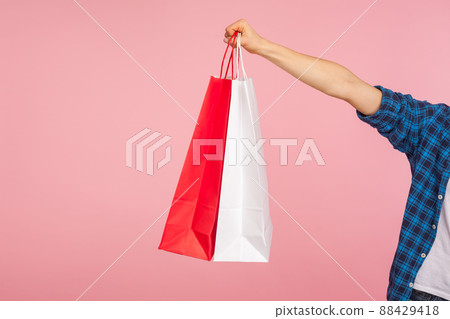 Closeup of woman hand in checkered shirt holding shopping bags, showing red and white packages, copy space for store advertising, promotional text. indoor studio shot isolated on pink background 88429418