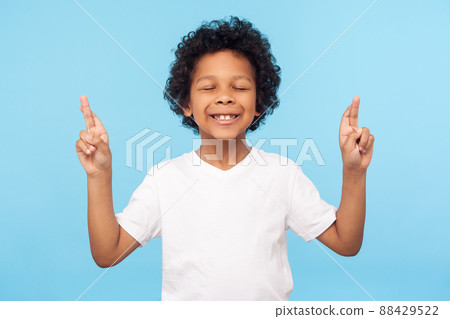 Childhood dreams and hopes. Portrait of adorable little boy standing with closed eyes and crossing fingers, wishing for good luck, waiting miracle. indoor studio shot isolated on blue background Childhood dreams and hopes. Portrait of adorable little boy standing with closed eyes and crossing fingers, wishing for good luck, waiting miracle. indoor studio shot isolated on blue background 88429522