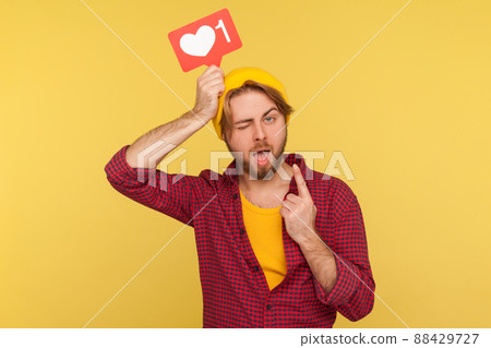 Optimistic hipster guy in checkered shirt holding social network Heart Like icon over head and raising finger with great idea gesture, winking at camera. studio shot isolated on yellow background 88429727