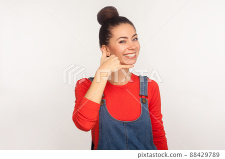 Phone conversation. Portrait of positive stylish pretty girl with hair bun in denim overalls showing call me gesture with hand shaped like telephone. indoor studio shot isolated on white background 88429789