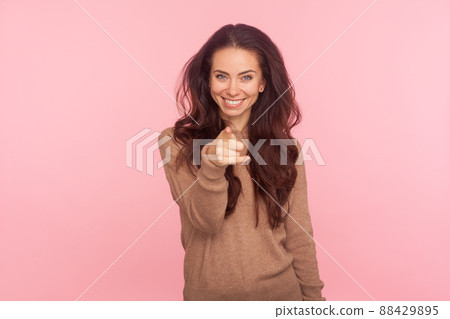 Hey you. Portrait of cheerful young woman with brunette wavy hair pointing to camera and looking with charming toothy smile, indicating finger, making choice. studio shot isolated on pink background 88429895