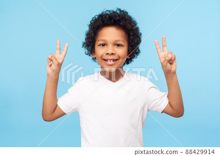 Portrait of happy and victorious little boy in T-shirt showing victory or peace gesture and smiling, doing v sign, number two, cute child enjoying win. indoor studio shot isolated on blue background Portrait of happy and victorious little boy in T-shirt showing victory or peace gesture and smiling, doing v sign, number two, cute child enjoying win. indoor studio shot isolated on blue background 88429914