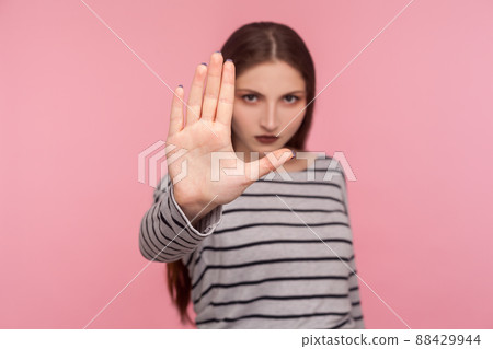 Portrait of strict serious woman in striped sweatshirt showing stop to camera, warning of danger, denying conflict with negative prohibition hand gesture. studio shot isolated on pink background Portrait of strict serious woman in striped sweatshirt showing stop to camera, warning of danger, denying conflict with negative prohibition hand gesture. studio shot isolated on pink background 88429944