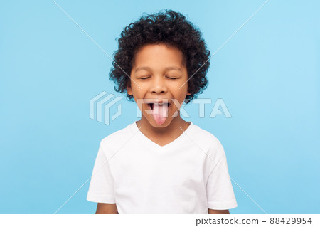 Portrait of funny naughty little boy with curly hair in T-shirt sticking out tongue and keeping eyes closed, disobedient child foolishing with cute derisive expression. studio shot blue background 88429954