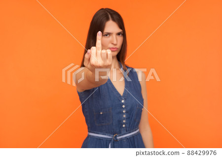 Impolite annoyed brunette woman in denim dress showing middle finger to camera, expressing protest hate and disrespect, gesturing fuck off rude symbol. indoor studio shot isolated on orange background 88429976