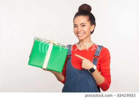 Look at gift. Portrait of positive joyful pretty girl with hair bun in denim overalls pointing at wrapped present box and smiling, congratulating on holidays, offering bonus. studio shot, isolated Look at gift. Portrait of positive joyful pretty girl with hair bun in denim overalls pointing at wrapped present box and smiling, congratulating on holidays, offering bonus. studio shot, isolated 88429983