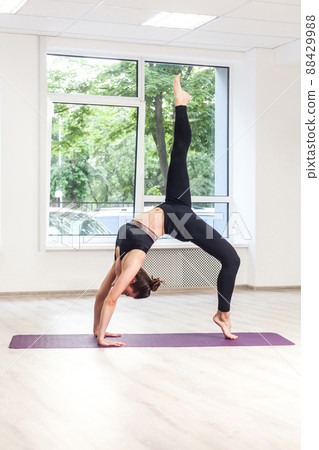 Full lenght side view portrait of sporty beautiful young adult woman in black sportswear practicing yoga, standing in bridge exercise, one legged wheel pose, working out, window background, indoor 88429988