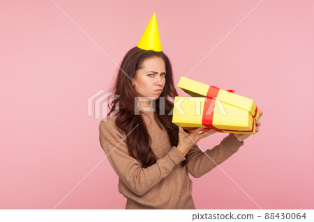 Portrait of funny displeased young woman with party cone on head unboxing gift and looking disappointed, upset about birthday present, delivery mistake. indoor studio shot isolated on pink background Portrait of funny displeased young woman with party cone on head unboxing gift and looking disappointed, upset about birthday present, delivery mistake. indoor studio shot isolated on pink background 88430064