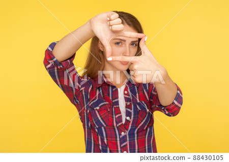 Portrait of beautiful ginger girl in checkered shirt looking at camera through fingers, photo frame gesture, focusing and pretending to make picture. indoor studio shot isolated on yellow background Portrait of beautiful ginger girl in checkered shirt looking at camera through fingers, photo frame gesture, focusing and pretending to make picture. indoor studio shot isolated on yellow background 88430105