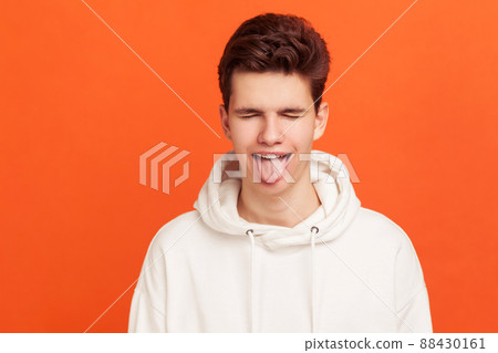 Mischievous boy in casual white hoodie closing eyes and showing tongue. Naughty teenager fooling around. Infantilism. Indoor studio shot isolated on orange background 88430161