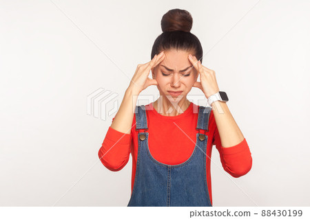 Headache. Portrait of tired depressed upset girl with hair bun in denim overalls feeling unhealthy, suffering migraine and tension, meningitis, flu symptom. studio shot isolated on white background Headache. Portrait of tired depressed upset girl with hair bun in denim overalls feeling unhealthy, suffering migraine and tension, meningitis, flu symptom. studio shot isolated on white background 88430199