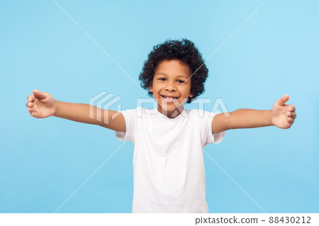Come here, free hugs. Portrait of lovely good-natured little boy with curly hair in white T-shirt smiling excitedly and holding hands wide open to embrace, greeting. indoor studio shot blue background 88430212