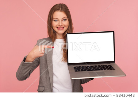 Portrait of beautiful elegant brunette businesswoman in suit jacket pointing at mock up display, holding laptop with blank screen for advertising. indoor studio shot isolated on pink background 88430448