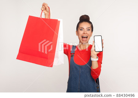 Portrait of excited amazed shopper girl in denim overalls holding shopping bags and cell phone, advertising application for online ordering, purchase and delivery of goods. studio shot, isolated Portrait of excited amazed shopper girl in denim overalls holding shopping bags and cell phone, advertising application for online ordering, purchase and delivery of goods. studio shot, isolated 88430496