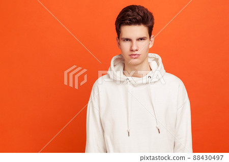 Serious young man in light sweater with hood looking at camera, confident teenager ready for adult life. Indoor studio shot isolated on orange background Serious young man in light sweater with hood looking at camera, confident teenager ready for adult life. Indoor studio shot isolated on orange background 88430497