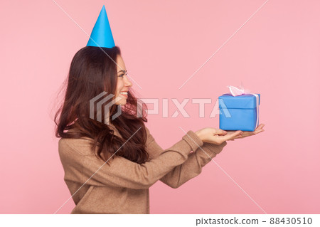 Take this present. Side view of happy generous nice young woman wearing party cone on head holding gift box, congratulating and offering birthday present. studio shot isolated on pink background 88430510