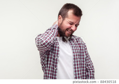 Portrait of exhausted bearded man in casual plaid shirt massaging aching neck with expression of unbearable suffer, having spine problems, back pain. indoor studio shot isolated on white background Portrait of exhausted bearded man in casual plaid shirt massaging aching neck with expression of unbearable suffer, having spine problems, back pain. indoor studio shot isolated on white background 88430518
