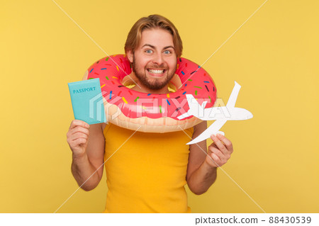 Excited optimistic funny tourist guy in undershirt standing with pink donut rubber ring, holding passport document and airplane mockup, rejoicing travel tour. studio shot isolated on yellow background Excited optimistic funny tourist guy in undershirt standing with pink donut rubber ring, holding passport document and airplane mockup, rejoicing travel tour. studio shot isolated on yellow background 88430539