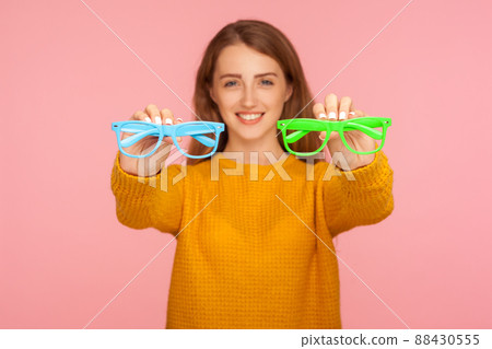 Optics and eyesight. Portrait of cheerful ginger girl in sweater holding green and blue eyeglasses and smiling at camera, recommending spectacles. indoor studio shot isolated on pink background Optics and eyesight. Portrait of cheerful ginger girl in sweater holding green and blue eyeglasses and smiling at camera, recommending spectacles. indoor studio shot isolated on pink background 88430555