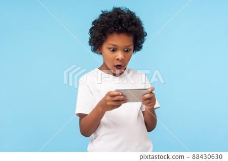Amazed surprised funny little boy with curly hair using mobile phone with shocked expression, astonished kid playing video game on cellphone, children entertainment. indoor studio shot blue background 88430630