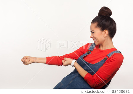 Portrait of strong ambitious girl with hair bun in denim overalls pretending to pull invisible rope, concept of hard working, striving and efforts to achievements. indoor studio shot, white background Portrait of strong ambitious girl with hair bun in denim overalls pretending to pull invisible rope, concept of hard working, striving and efforts to achievements. indoor studio shot, white background 88430681
