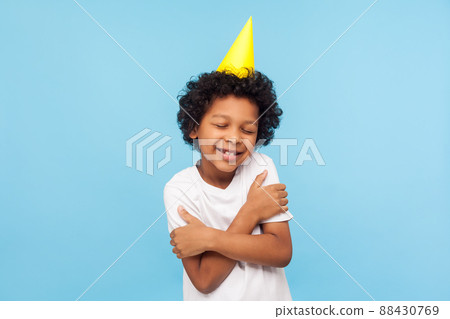 I love and congratulate myself. Portrait of amazing lovely kid with funny party cone embracing himself and smiling from pleasure, happy to celebrate birthday alone. indoor studio shot blue background 88430769