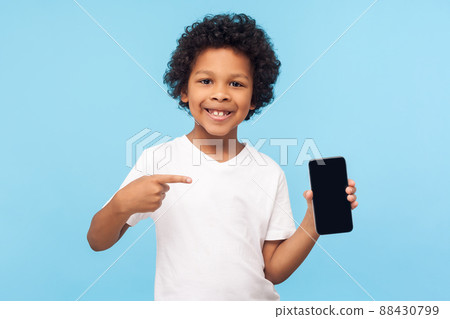 Mobile app for children. Happy excited cheerful little boy with curly hair in T-shirt pointing at cell phone and smiling at camera, showing telephone. indoor studio shot isolated on blue background 88430799
