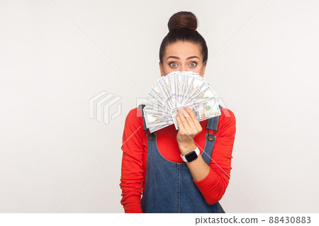 Portrait of astonished girl with hair bun in denim overalls peeking out of money dollars with scared surprised eyes, shocked by lottery win, big profit. indoor studio shot isolated on white background 88430883