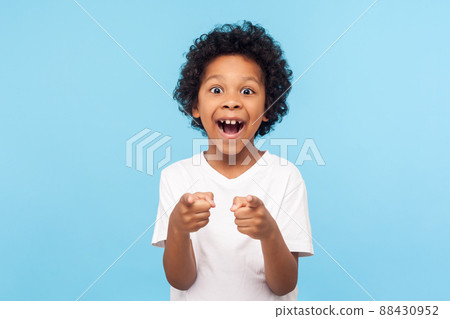 Hey you. Portrait of happy little boy with curly hair pointing finger to camera and laughing loudly with surprised face, teasing making fun of you. indoor studio shot isolated on blue background 88430952