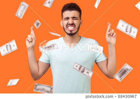 Hope to win money. Portrait of hopeful man standing, closed eyes and clenched teeth, crossing his fingers and eagerly wishing to be rich. money falling from top. indoor, isolated on orange background 88430967