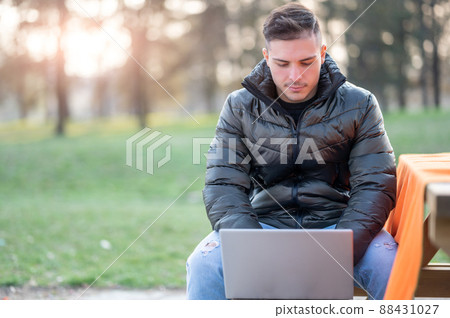 A attractive young office worker in casual wear, is focused.while typing on his laptop. I work remotely on a bench in the park. Remote working outdoors concept. 88431027