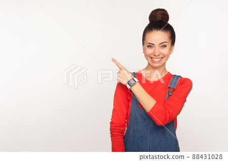 Attention to advertising. Portrait of happy stylish pretty girl with hair bun in denim overalls pointing aside to copy space, blank wall for commercial text. studio shot isolated on white background 88431028
