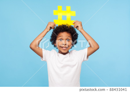 Funny excited surprised little boy with curly hair in T-shirt holding big yellow hashtag symbol over his head and looking with amazement, popular blogging, child content. studio shot n blue background 88431041