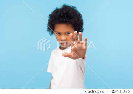 No, forbidden. Portrait of serious angry little boy with curly hair showing stop gesture, warning to go and looking aggressive, rejection denial concept. indoor studio shot isolated on blue background 88431250