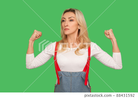 Women's rights, power to achieve goal. Portrait of self-confident energetic adult female in stylish overalls raising hands showing biceps, feeling independent and strong. indoor studio shot, isolated 88431592