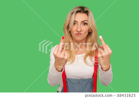 Portrait of angry impolite rude woman in denim overalls rejecting communication, showing double middle finger to express disrespect and hate, vulgar gesture. studio shot isolated on green background 88431625