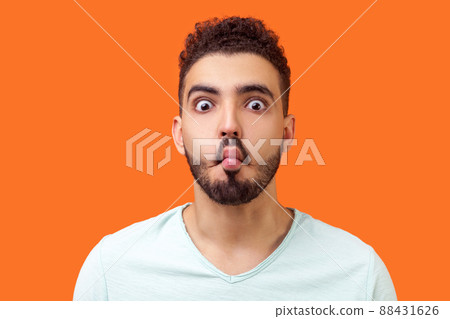 Portrait of funny silly brunette man with beard in casual white t-shirt looking at camera and making fish face with lips and big eyes, fooling around. indoor studio shot isolated on orange background 88431626