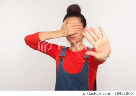 No, don't want to look. Portrait of displeased girl with hair bun in denim overalls covering eyes with hand and showing stop gesture, avoiding to watch. indoor studio shot isolated on white background 88431800