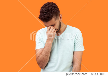 Portrait of depressed lonely brunette man with beard in casual white t-shirt holding head down, hiding his face in hand and crying, feeling desperate. indoor studio shot isolated on orange background 88431806
