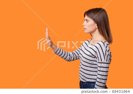 Side view portrait of worried young woman with brown hair in long sleeve striped shirt standing, raising arm showing stop gesture, warning sign. indoor studio shot isolated on orange background Side view portrait of worried young woman with brown hair in long sleeve striped shirt standing, raising arm showing stop gesture, warning sign. indoor studio shot isolated on orange background 88431864