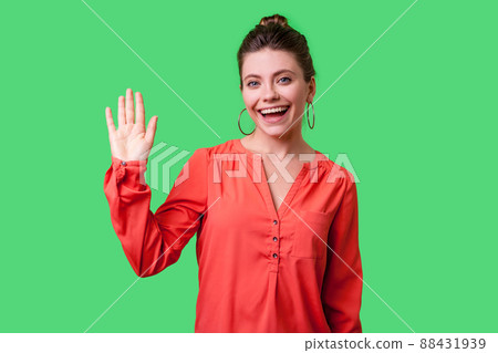 Hello. Portrait of charming woman with bun hairstyle, big earrings and in red blouse standing, waving hand and saying Hi to camera with toothy smile. indoor studio shot isolated on green background Hello. Portrait of charming woman with bun hairstyle, big earrings and in red blouse standing, waving hand and saying Hi to camera with toothy smile. indoor studio shot isolated on green background 88431939