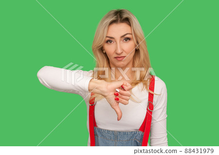 Dislike, bad rating. Portrait of dissatisfied displeased adult woman in stylish denim overalls showing thumbs down gesture to critisize awful service. indoor studio shot isolated on green background Dislike, bad rating. Portrait of dissatisfied displeased adult woman in stylish denim overalls showing thumbs down gesture to critisize awful service. indoor studio shot isolated on green background 88431979