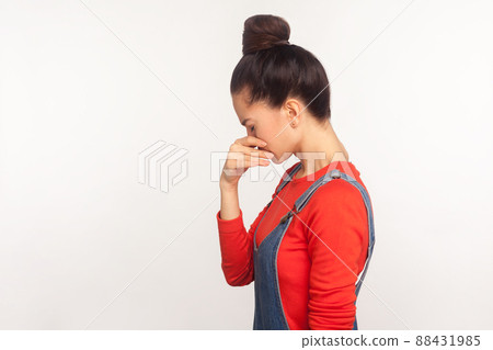Sadness and depression. Side view of unhappy girl with hair bun in denim overalls bowing her head and crying, experiencing desperate sorrowful feelings. indoor studio shot isolated on white background Sadness and depression. Side view of unhappy girl with hair bun in denim overalls bowing her head and crying, experiencing desperate sorrowful feelings. indoor studio shot isolated on white background 88431985